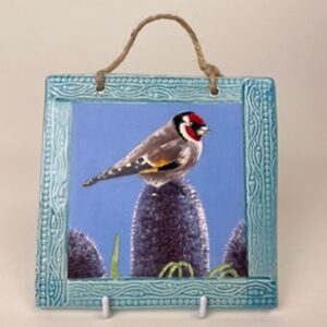 A handmade earthenware hanging wall tile. This tile design of a gold finch on a thistle symbolises light and sanctity. 