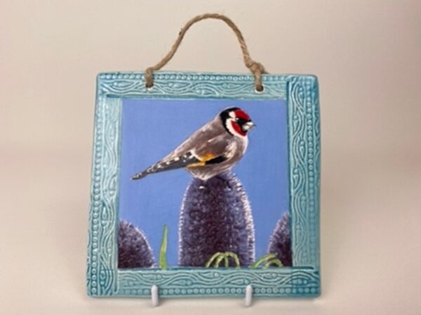 A handmade earthenware hanging wall tile. This tile design of a gold finch on a thistle symbolises light and sanctity. 